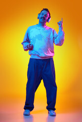 Energetic, smiling man in sweatshirt and jeans listening to music in headphone, gesturing and dancing isolated on gradient yellows orange background in neon. Human emotions, casual fashion concept
