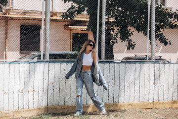 Beautiful blonde model, fashionable in jeans and a denim jacket, poses in the city against the background of a sports field grid