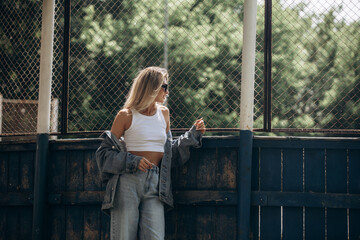 Beautiful blonde model, fashionable in jeans and a denim jacket, poses in the city against the background of a sports field grid