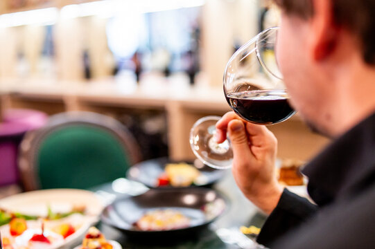 Person holding a glass of red wine, with a blurred dining setting and various dishes in the background. The image evokes a sense of sophistication and enjoyment