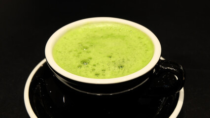 Closeup of a cup of matcha green tea drink, with some froth on the top. With a black cup and a black background.