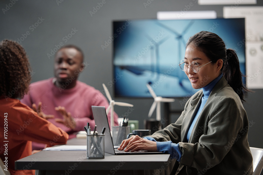 Poster Young smiling businesswoman in formalwear sitting by desk with windmill models and networking against colleagues discussing working points