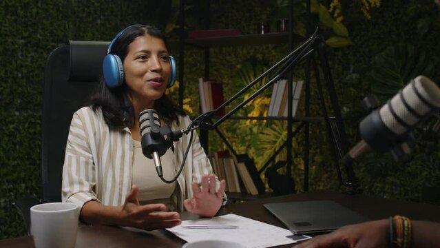Over the shoulder shot of you Latin woman in headphones smiling and talking in mic while hosting podcast interview with guest in recording studio