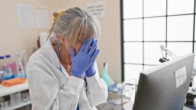 Mature woman scientist feeling stressed in a laboratory setting, with computers and lab equipment.