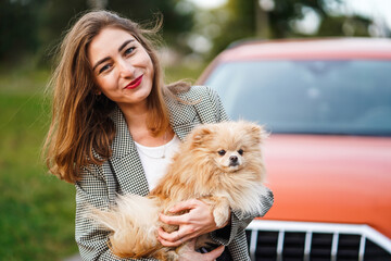 Smiling Woman Holding A Fluffy Pomeranian Dog Near A Red Car In The Park