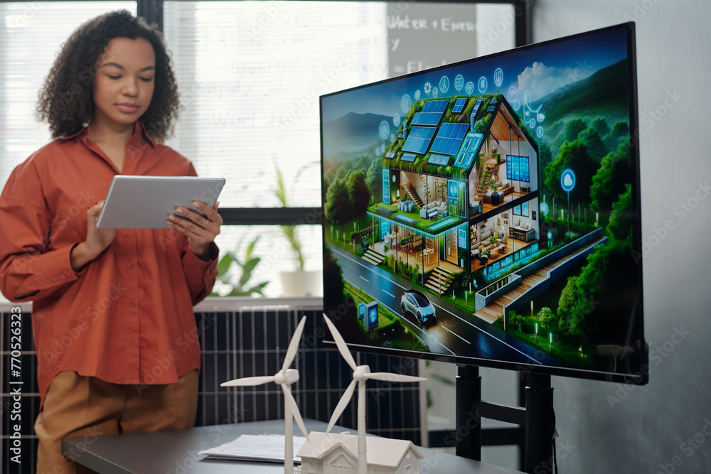 Poster Screen with new architectural project and two models of windmill standing on desk against young female employee making presentation