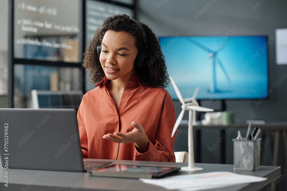 Poster young smiling female employee explaining something to person during communication in video chat whil