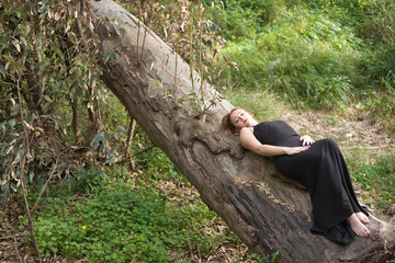 Young woman, blonde and beautiful, wearing a black dress, with angelic look, lying on the trunk of a large tree, in the middle of nature. Concept nature, peace, tranquility, trees.