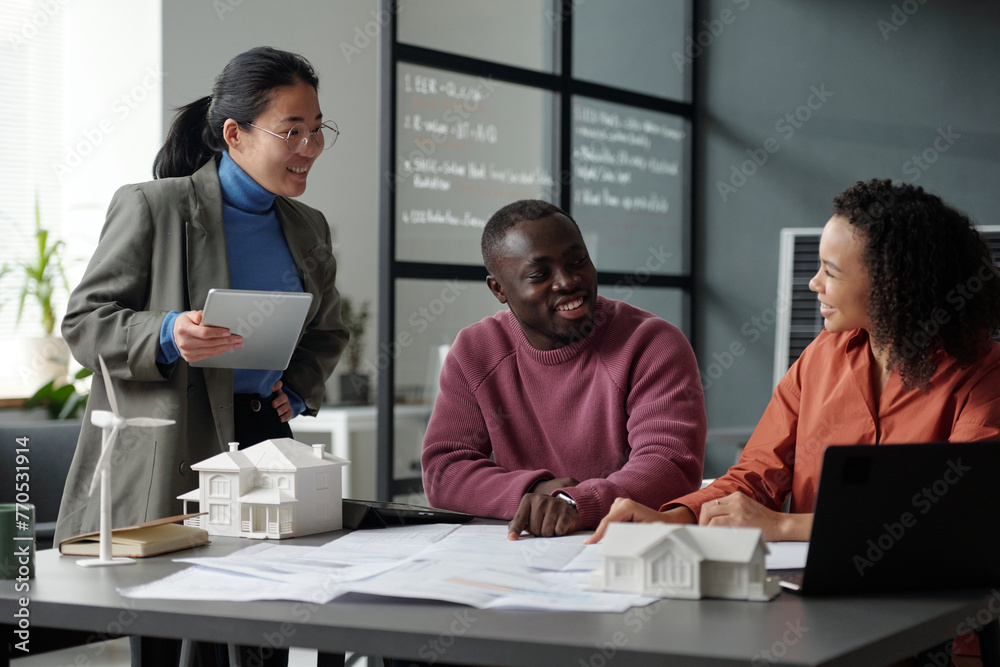 Aufkleber Three happy young intercultural architects discussing new project while one of them pointing at unrolled blueprint with sketch of house
