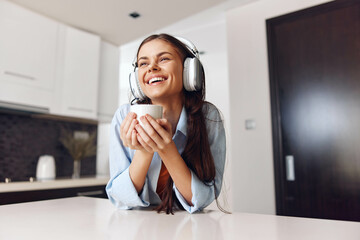 Naklejka premium Happy woman enjoying music and coffee in cozy kitchen at home, listening to her favorite tunes through headphones