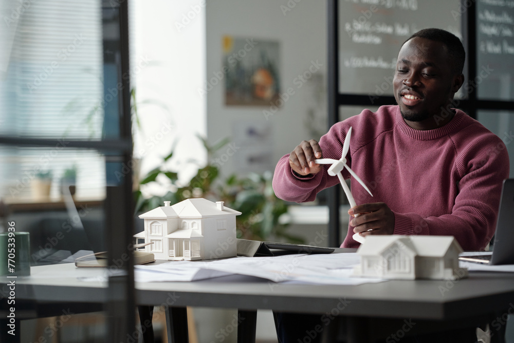 Poster Young smiling male architect rotating turbine of windmill model while sitting by workplace with unrolled blueprint and house mockup