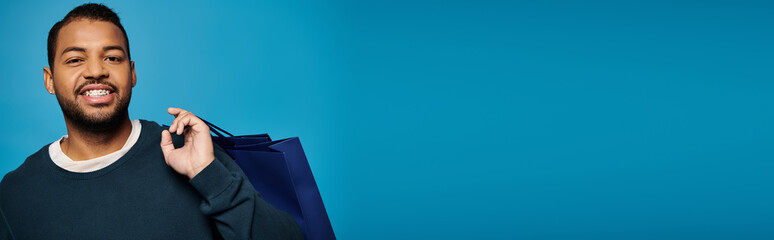 banner of happy african american young man with shopping bag over shoulder on blue background