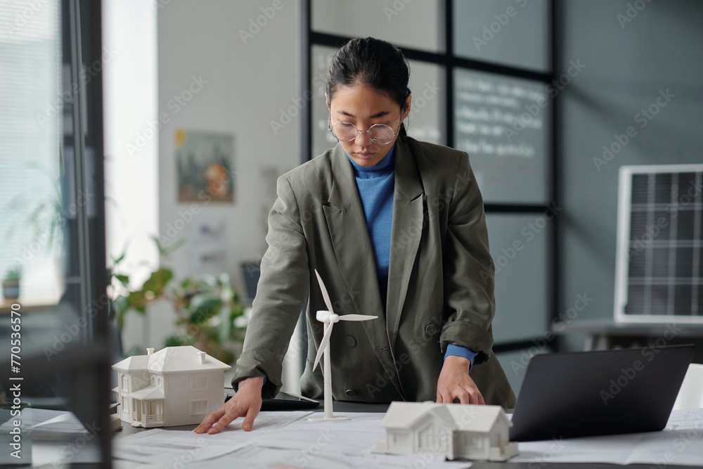 Poster young asian female architect bending over workplace with blueprint and looking at sketch while worki