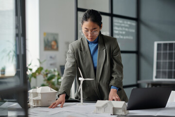 Young Asian female architect bending over workplace with blueprint and looking at sketch while...