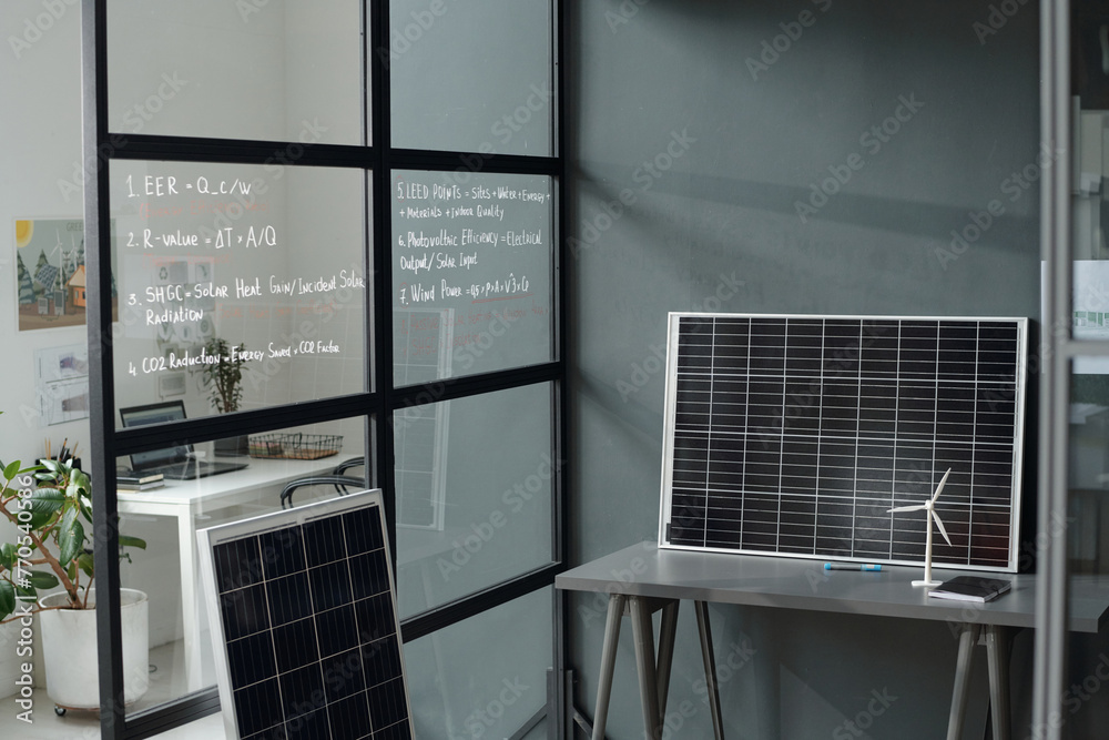 Poster grey desk with models of solar panel and windmill standing by wall of spacious modern office with wo