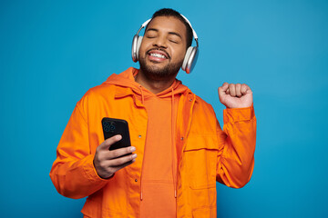 cheerful african american man in orange outfit and headphones dancing with smartphone