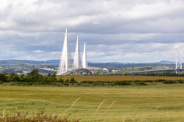 Queensferry Crossing white suspension bridge in green grass farmland across water, sea Firth of Forth near Edinburgh, Scotland, Europe