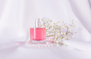 Glass bottle of pink perfume on white satin background with gypsophila branch and pearl beads. The concept of natural floral scents. Front view.