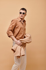 Fashion shot of man in beige shirt with sunglasses and holding jacket in hand on beige backdrop