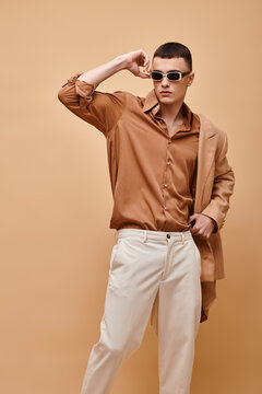Classy man in beige jacket on shoulder, shirt, pants and sunglasses posing on beige background