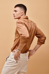 Back view portrait of stylish man in beige shirt with hand on pants on beige background