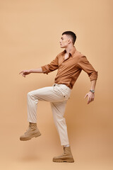 Fototapeta na wymiar Side view photo of young man in beige shirt, pants and boots posing on beige background