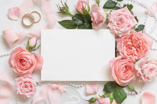 Blank card near pink roses, wedding rings and silk ribbons top view, wedding mockup