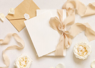 Cards tied with a beige silk ribbon near cream roses and envelope on white table top view, mockup
