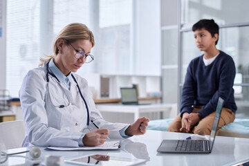 Portrait of woman doctor wearing glasses taking notes with child sitting on cot in background copy...