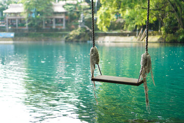 A traditional swing located right above the blue lake and surrounded by shady green trees and goldfish swimming beneath it