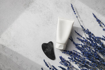 Top view of dark guasha stone and white creme tube, fresh aromatic lavender flowers on white background.Natural light and shadows. Wellness being