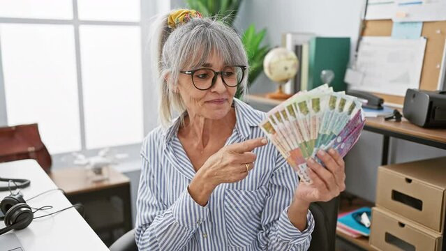 Joyful middle age woman with grey hair ecstatically pointing at hong kong dollars banknotes in hand at the office, beaming with hope and positivity