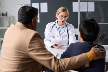 Portrait of mature woman doctor consulting Middle Eastern father and son in sitting across table in pediatric clinic