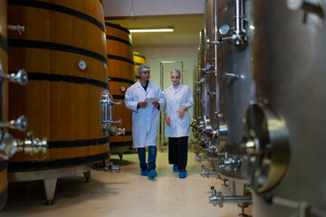 Winery technicians with clipboard and wine glass in hand walk between large oak barrels and...