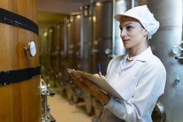 A focused female quality control specialist in a white coat inspects wine barrels in a fermentation...