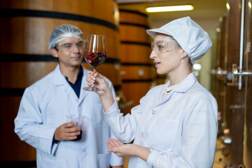 In a winery's aging room, a focused female wine expert evaluates the aroma of a wine sample while a male colleague observes.