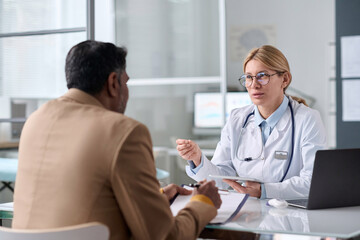 Portrait of smiling female doctor consulting Middle Eastern man sitting across table in medical clinic