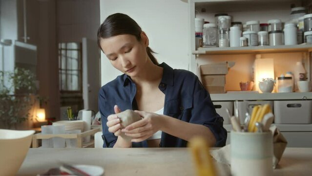 young Asian female artisan sits at table in ceramic studio. freelance ceramist shaping and forming clay for pottery.