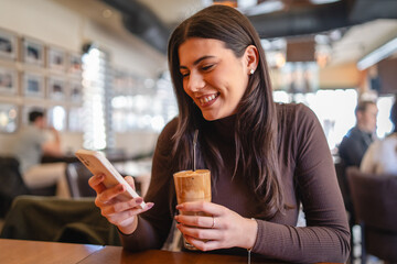 A one young happy girl or woman is drinking cold coffee in cafe or restaurant while using her phone...