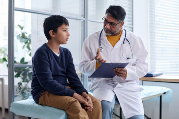 Portrait of Middle Eastern doctor talking to young boy during exam in medical clinic both sitting...