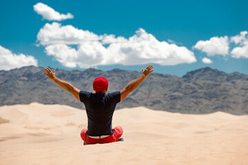 Happy tourist in red cap is sitting with open arms against sand dunes, mountains and blue sky. Travel in desert concept