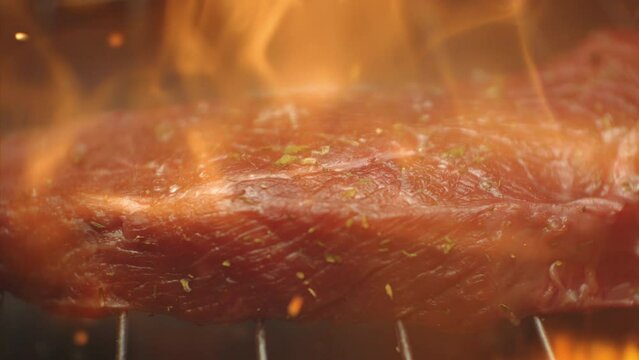 Grilled meat steak on stainless grill depot with fire on dark background. Preparing tasty raw red beef steak on a grill with flame. Food and cuisine concept. Cooking process in slow motion 1000fps