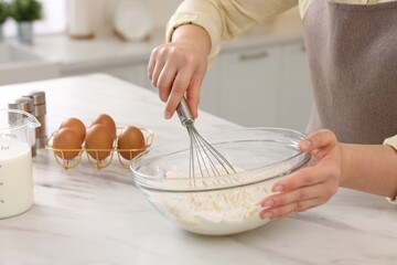 Woman making dough with whisk in bowl at light marble table, closeup