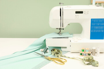 Modern sewing machine with cloth and craft accessories on white table near light green wall