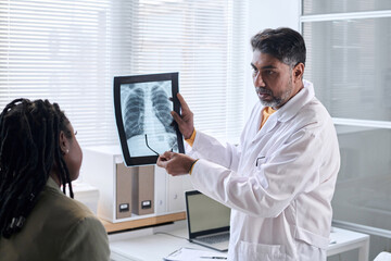 Waist up portrait of Middle Eastern doctor pointing at chest x ray and explaining image to patient in clinic copy space