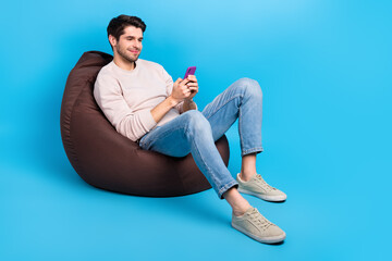 Full size photo of clever positive man wear sweatshirt denim pants sit on bean bag look at smartphone isolated on blue color background