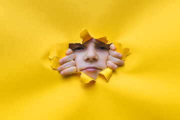 A little phlegmatic girl looks into a torn hole in yellow paper. The concept of espionage, childish curiosity, peeping, indifference to what is seen. Copy space. - obrazy, fototapety, plakaty