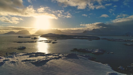 Sunset above Antarctica snow island, front aerial view. Sun reflection on ocean water in Antarctic polar summer. Beautiful sky horizon landscape. Drone forward flight