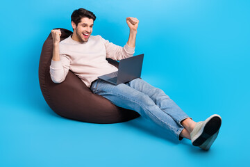 Full size photo of crazy satisfied man wear sweatshirt denim pants sit on bean bag win bet scream yes isolated on blue color background