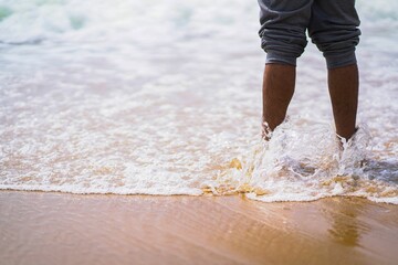 Legs of a person standing on the sandy shoreline against the lapping waves